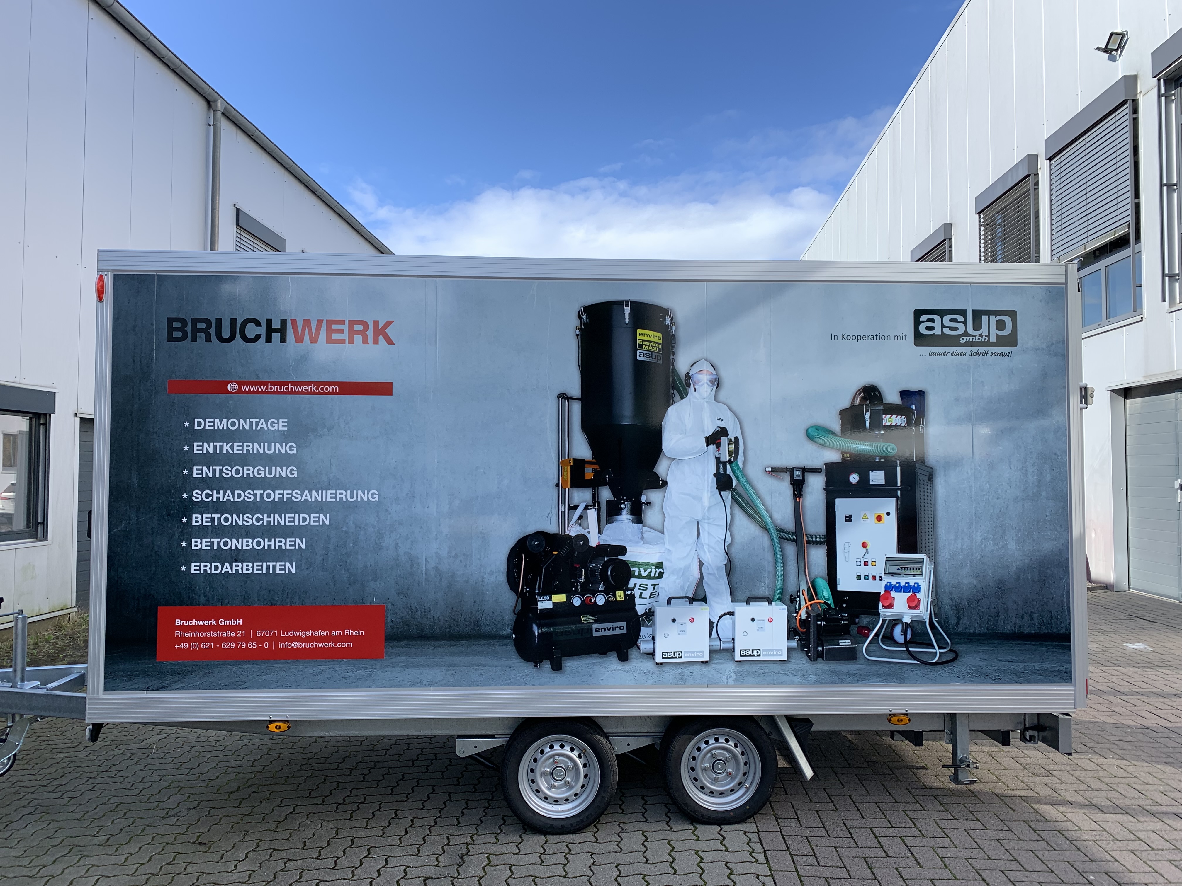 Das Bild zeigt einen Anhänger mit dem Logo "BRUCHWERK" und den Dienstleistungen: Demontage, Entkernung, Entsorgung, Schadstoffsanierung, Betonschneiden, Betonbohren und Erdarbeiten. Neben einem Arbeiter in Schutzkleidung sind verschiedene Maschinen und Geräte abgebildet.