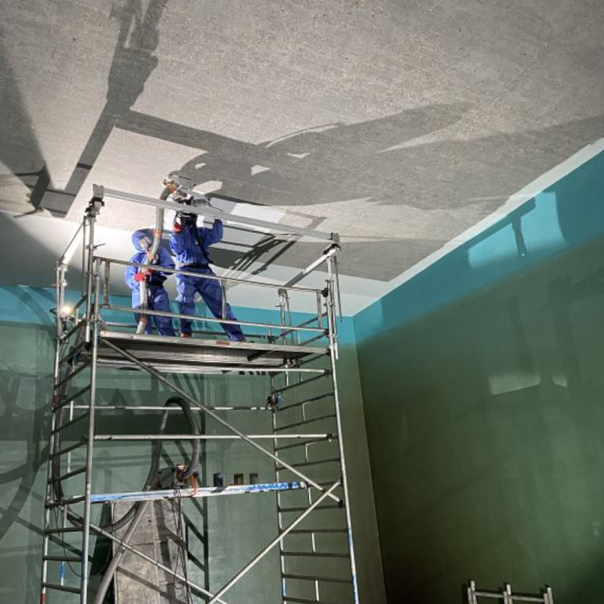 Zwei Arbeiter stehen auf einem Gerüst in einem Raum und fügen Material an die Decke hinzu. Einer von ihnen hält ein Werkzeug, während der andere unterstützt. Die Wände sind in verschiedenen Farbtönen gestrichen, und das Gerüst ist aus Metall.