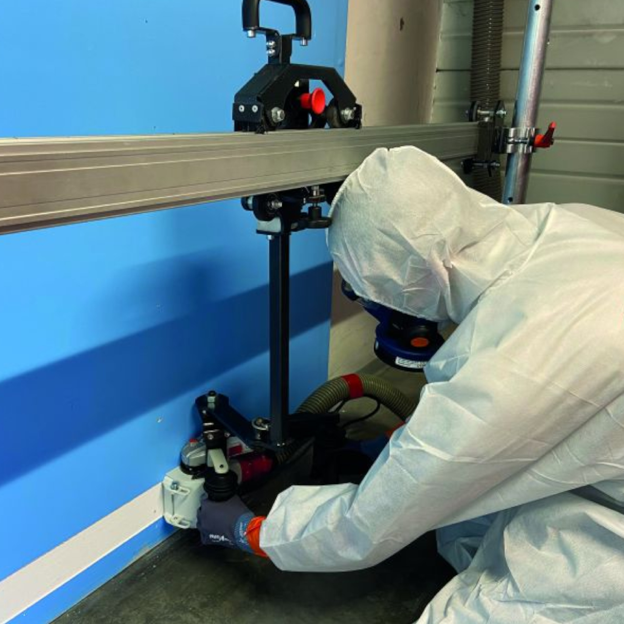 Ein Arbeiter in Schutzkleidung montiert ein Gerät an einer Wand. Im Hintergrund ist eine blaue Wand zu sehen. Der Arbeiter hält ein Werkzeug, das mit einer Schiene verbunden ist, während ein Staubsaugerschlauch sichtbar ist.