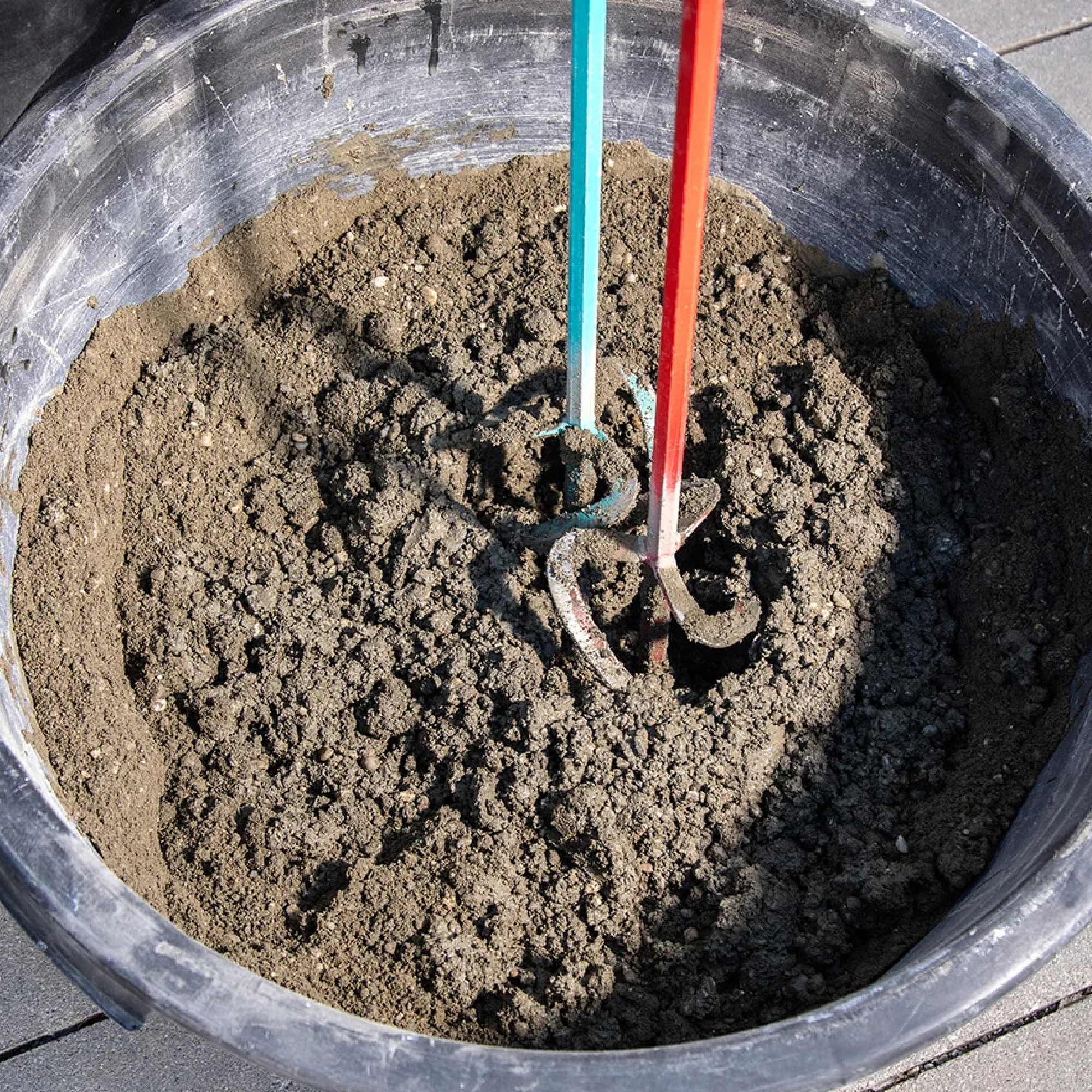 Ein schwarzer Eimer mit Erde, in dem zwei farbige Gartenwerkzeuge (rot und blau) stecken. Die Werkzeuge haben drei Zinken und sind teilweise im Erdreich verborgen. Die Szenerie vermittelt den Eindruck einer Gartenarbeit oder Vorbereitung für das Pflanzen.