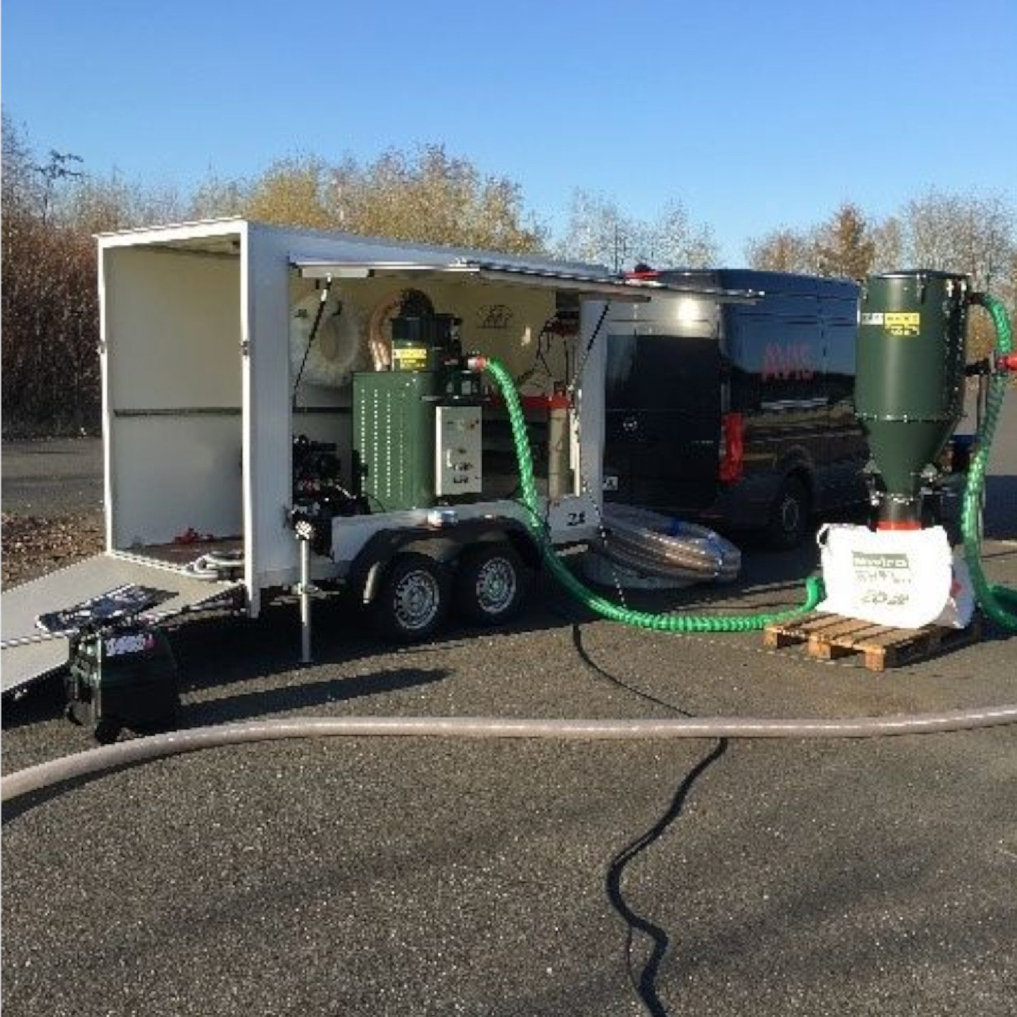 Eine mobile Maschinenanlage auf einem Anhänger, ausgestattet mit einem grünen Gerät und Schläuchen. Der Anhänger ist geöffnet, und im Hintergrund steht ein schwarzer Transporter. Der Standort wirkt industriell, umgeben von Bäumen und flachem Gelände.