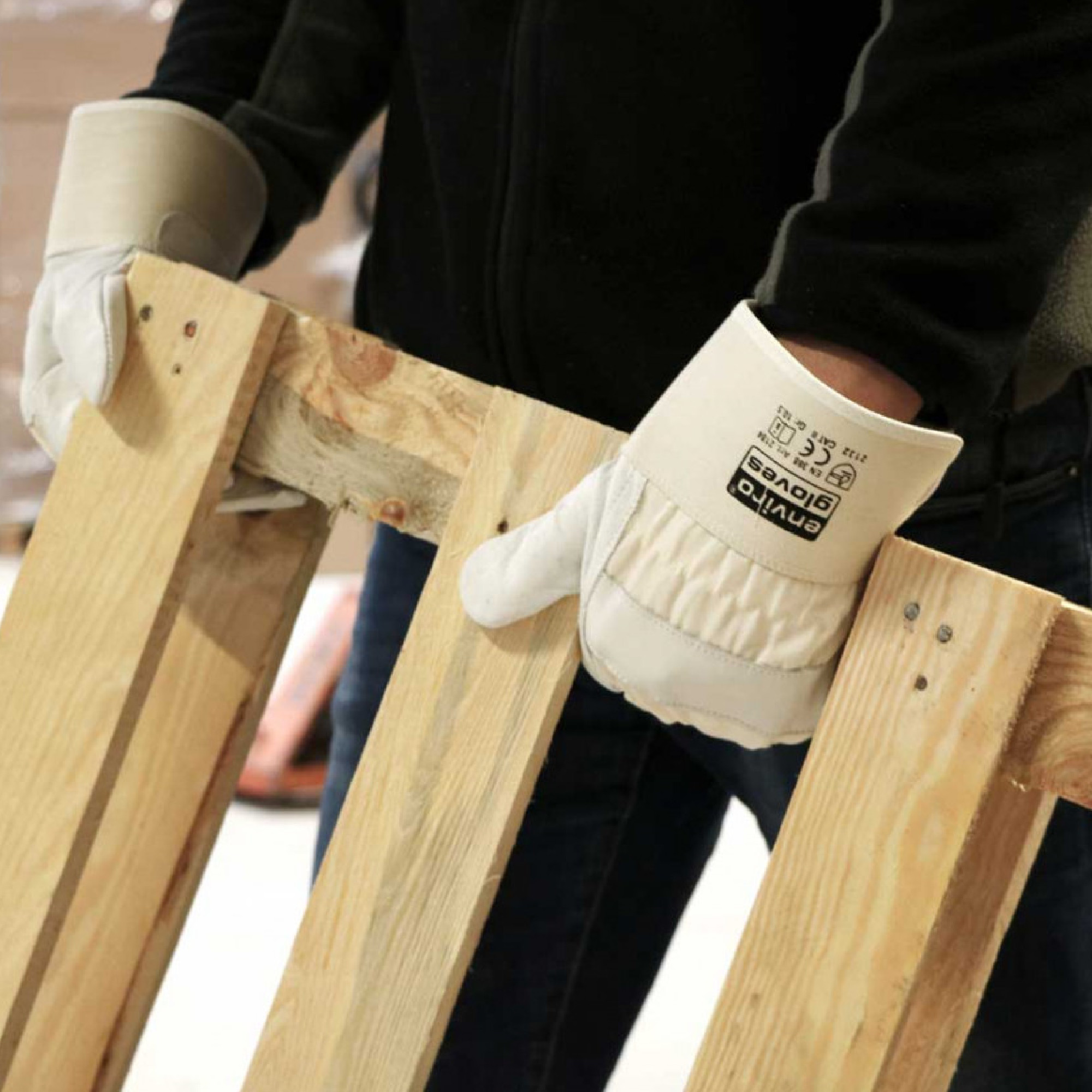 Eine Person trägt Arbeitshandschuhe und hält ein Holzstück oder eine Palette mit beiden Händen. Der Fokus liegt auf der Handhabung des Holzstücks, was auf eine handwerkliche Tätigkeit hinweist.
