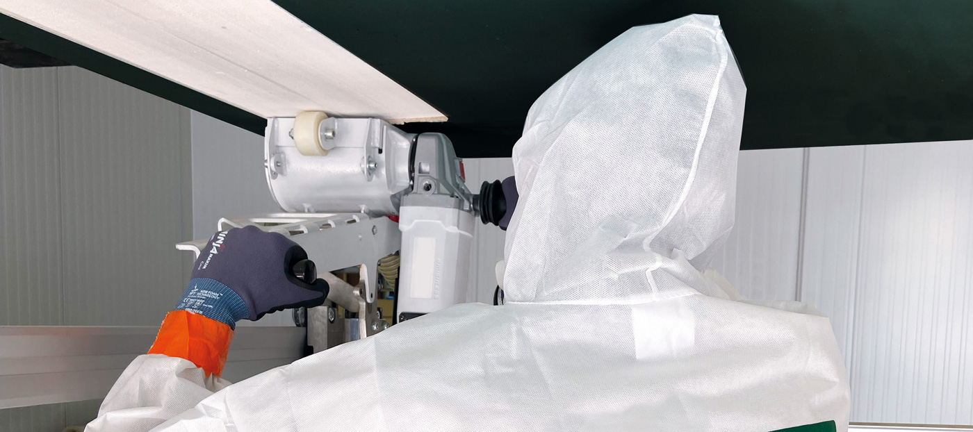 Eine Person in Schutzkleidung arbeitet mit einem Werkzeug an einer Maschine. Der Fokus liegt auf der Hand, die das Werkzeug hält, während die Maschine teilweise sichtbar ist. Der Hintergrund zeigt eine industrielle Umgebung.