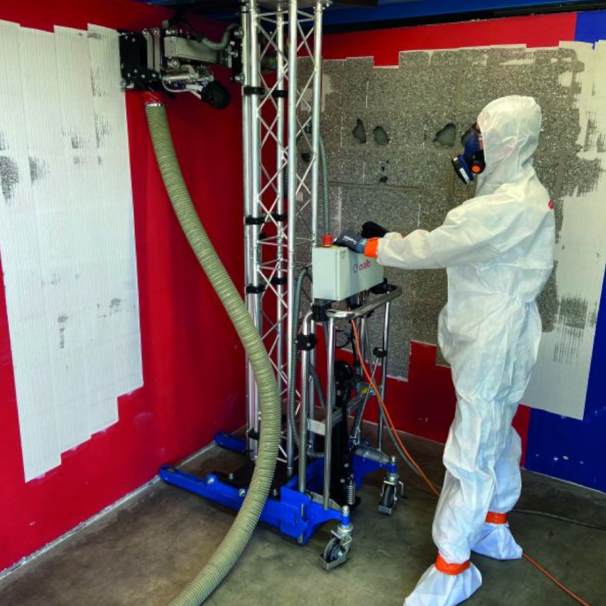 Ein Arbeiter in Schutzkleidung bedient ein Gerät zur Wandbearbeitung. Das Gerät steht vor einer roten Wand, die teilweise mit einem grauen Material bearbeitet wird. Ein Schlauch führt von der Maschine zur Wand.
