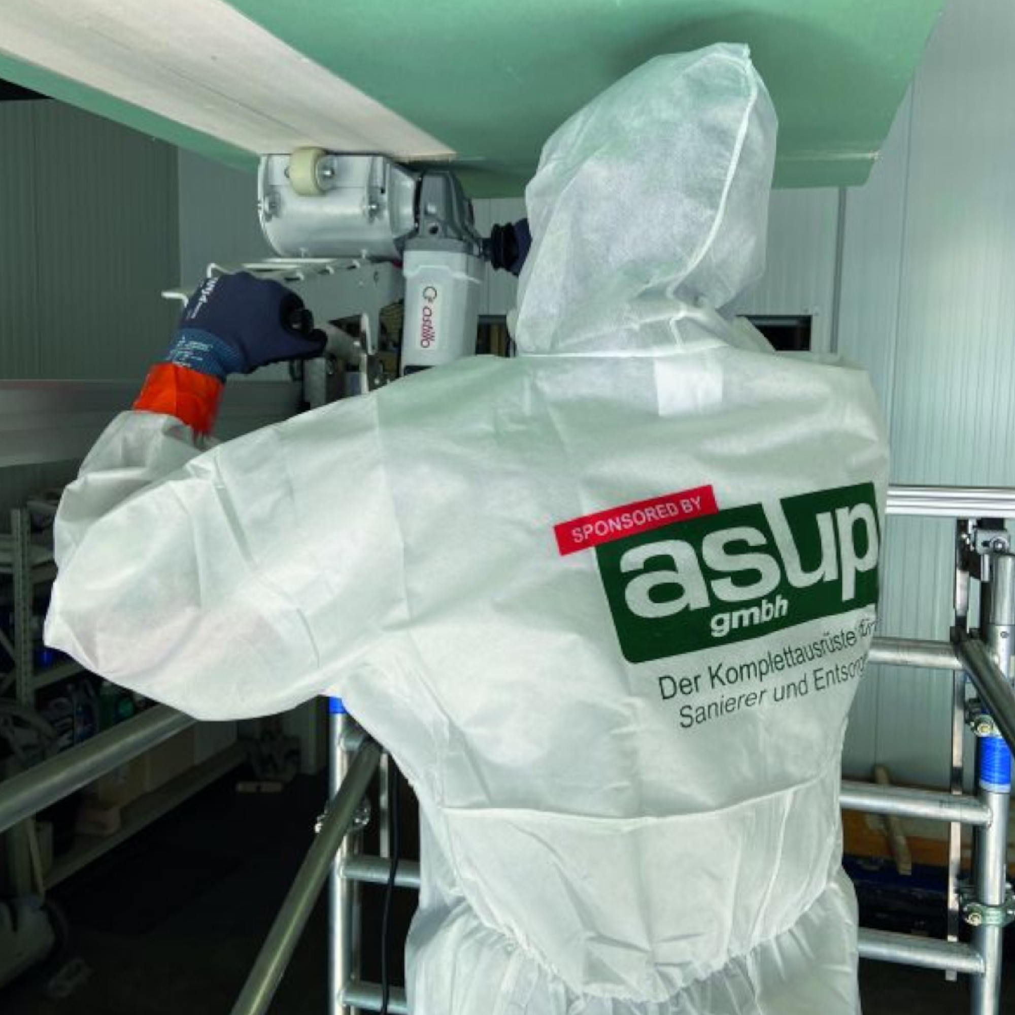 Ein Arbeiter in einem weißem Schutzanzug steht auf einer Leiter und bedient eine Maschine, die an der Decke montiert ist. Der Anzug trägt das Logo der Firma asup gmbh. Im Hintergrund sind einige Gegenstände und Gerüste sichtbar.
