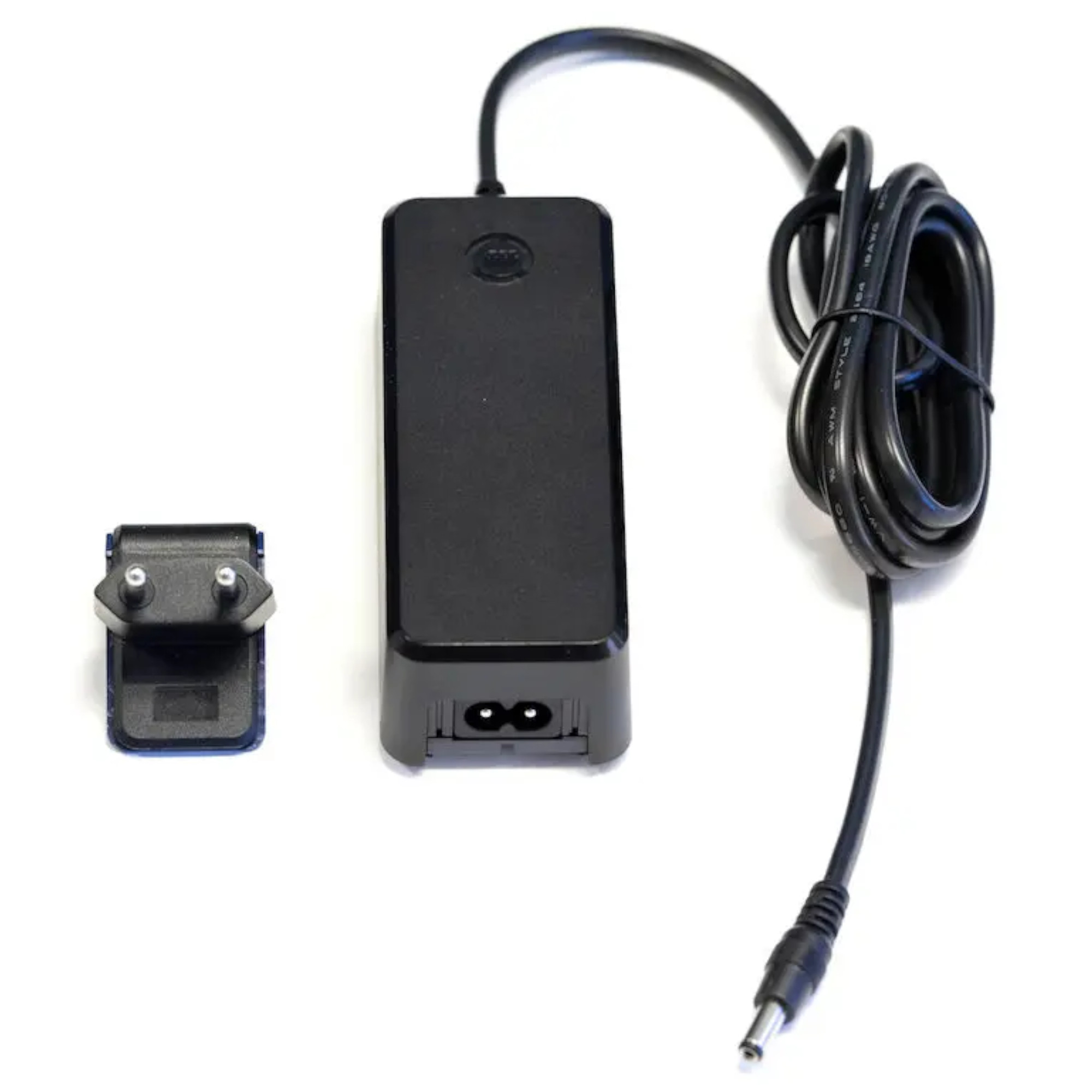 Ein schwarzes Netzteil mit einem Kabel und einem Stecker. Das Netzteil hat zwei Buchsen an der Unterseite und ist flach. Neben dem Netzteil liegt ein kleiner Adapter mit zwei Metallstiften. Das Kabel ist gewickelt.