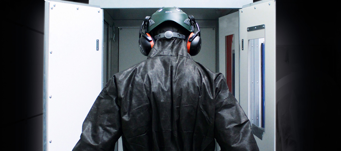 Eine Person mit einem schwarzen Schutzanzug und Gehörschutz steht vor offenen Türen eines Geräts oder Raums. Die Rückansicht zeigt den Vorbereitungsprozess für einen sicherheitsrelevanten Einsatz. Der Raum hat eine schlichte, technische Atmosphäre.