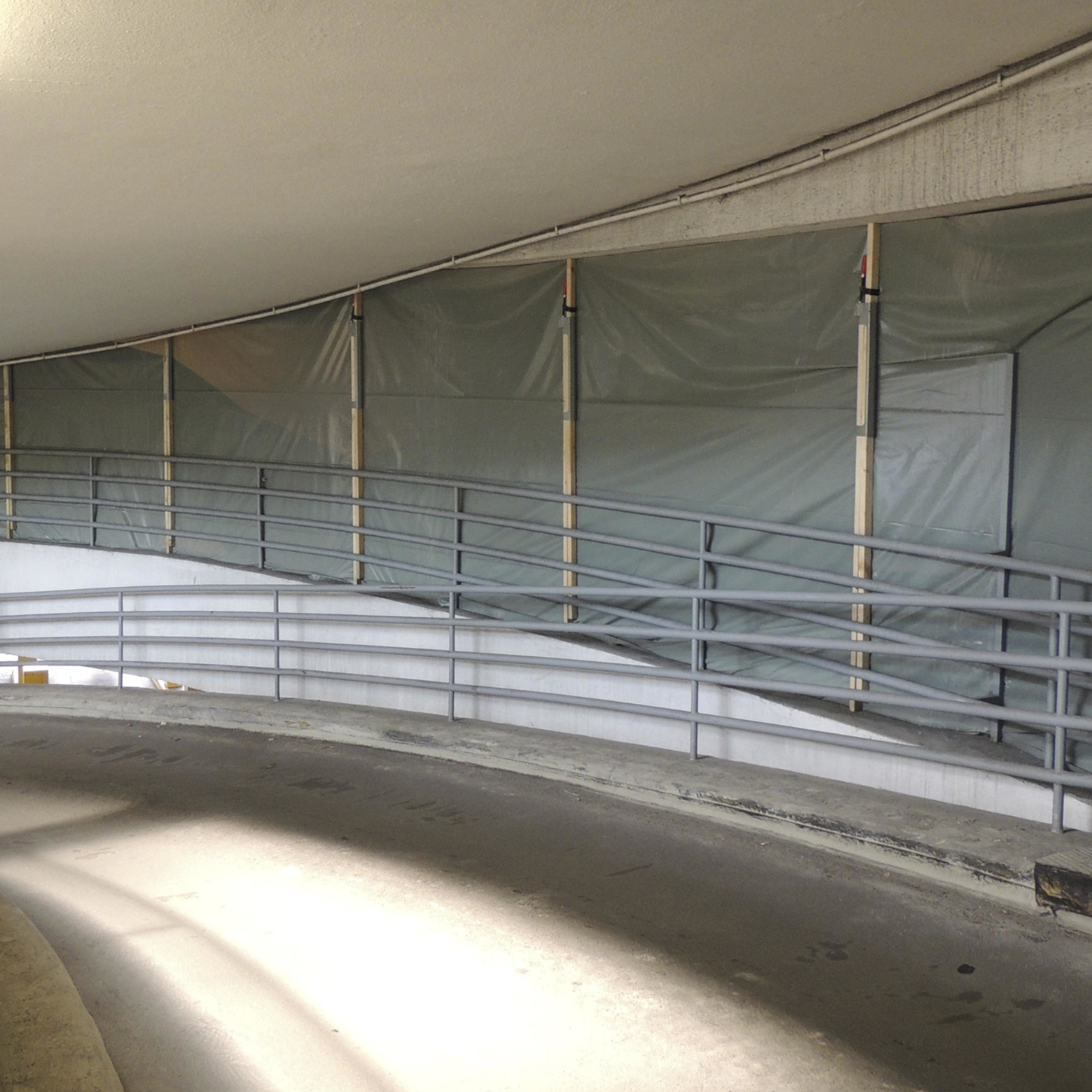 Ein gewundener Betonweg in einem Gebäude, an dem graue Wände mit Folie abgedeckt sind. Eine Metallgeländer verläuft entlang des Weges. Die Beleuchtung deutet auf eine helle Umgebung hin. Der Bereich scheint sich in der Nähe eines Eingangs oder Ausstiegs zu befinden.