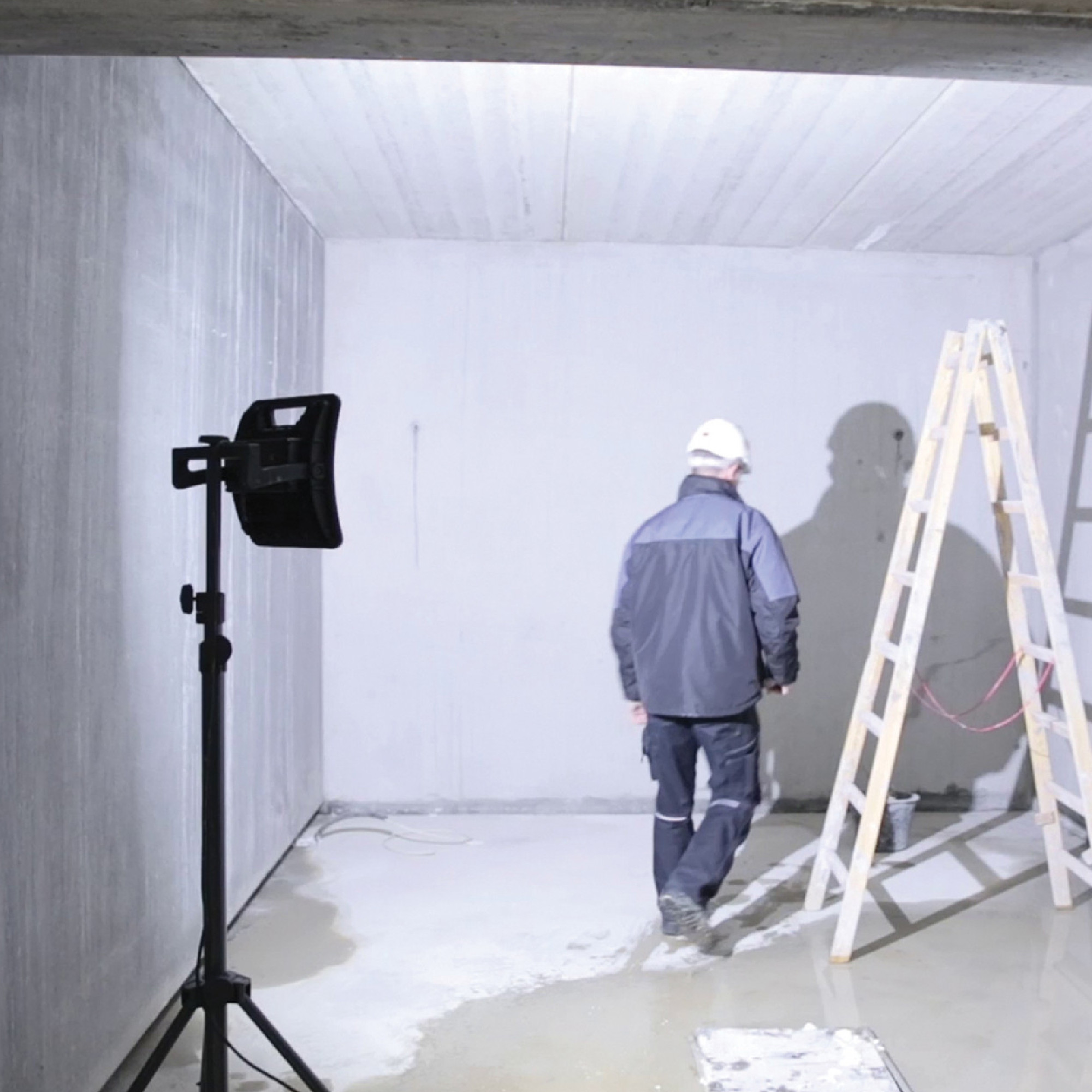 Ein Handwerker mit weißer Mütze und dunkler Kleidung bewegt sich in einem leeren, frisch gegossenen Raum. Im Hintergrund steht eine Holzleiter und ein Baustrahler beleuchtet die Szenerie. Die Wände sind aus Beton und der Boden ist teilweise nass.