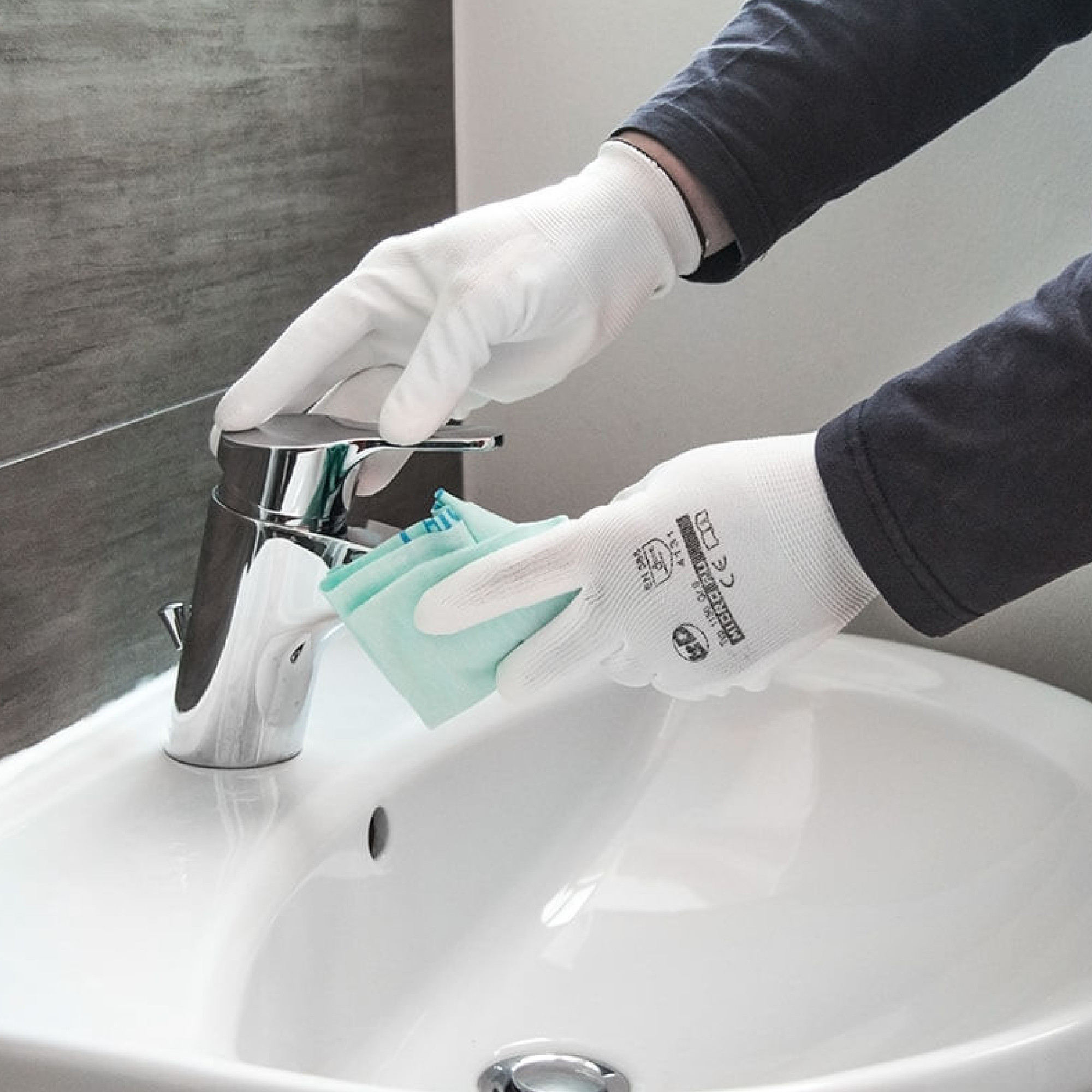 Eine Person mit weißen Handschuhen reinigt einen Wasserhahn in einem Waschbecken. Sie hält ein Reinigungstuch in der Hand und hat einen konzentrierten Gesichtsausdruck. Der Hintergrund ist hell und modern gestaltet.