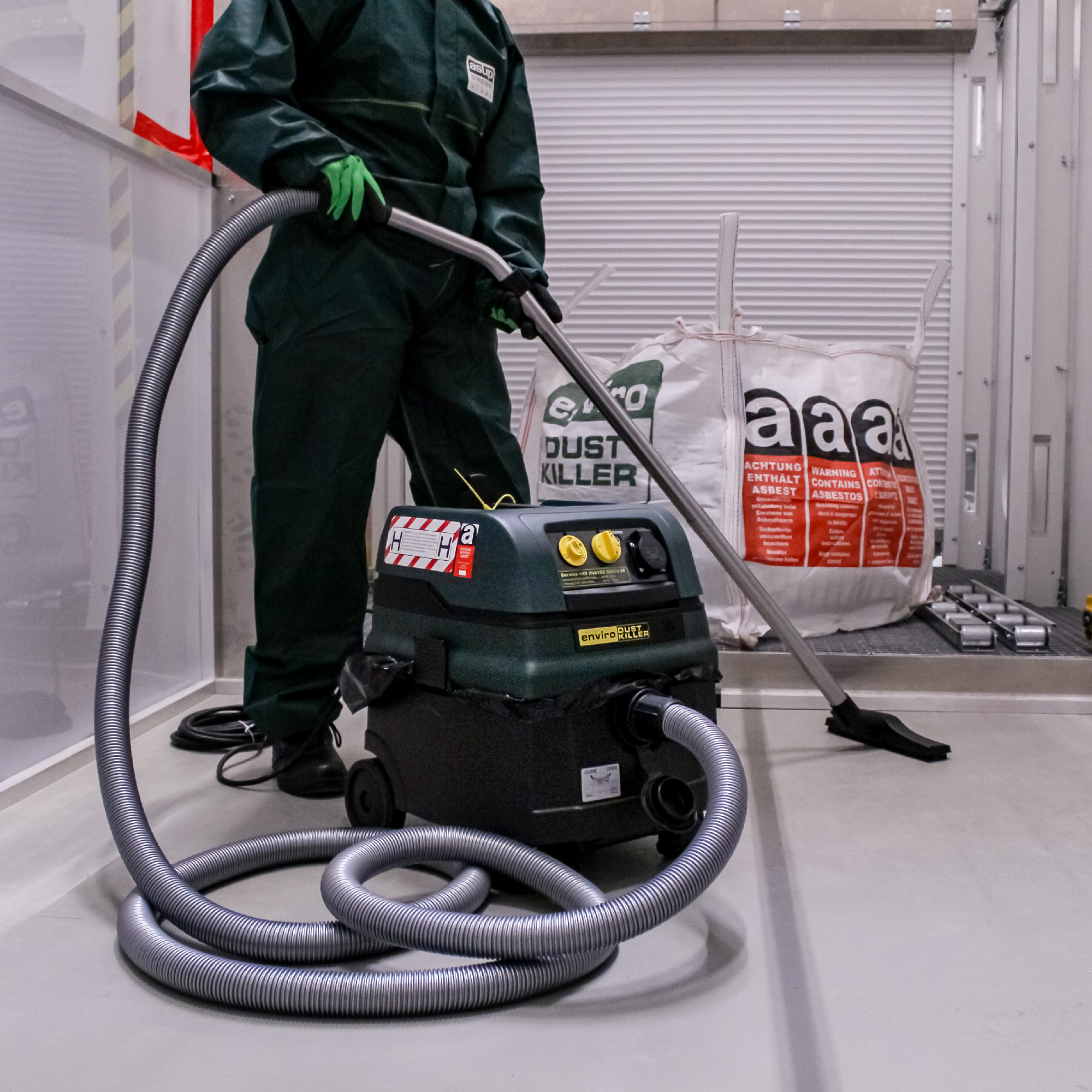 Person in grüner Schutzkleidung neben einem Staubsauger mit grauem Schlauchsystem; im Hintergrund Säcke mit „DUST KILLER“ und Asbest-Warnhinweisen