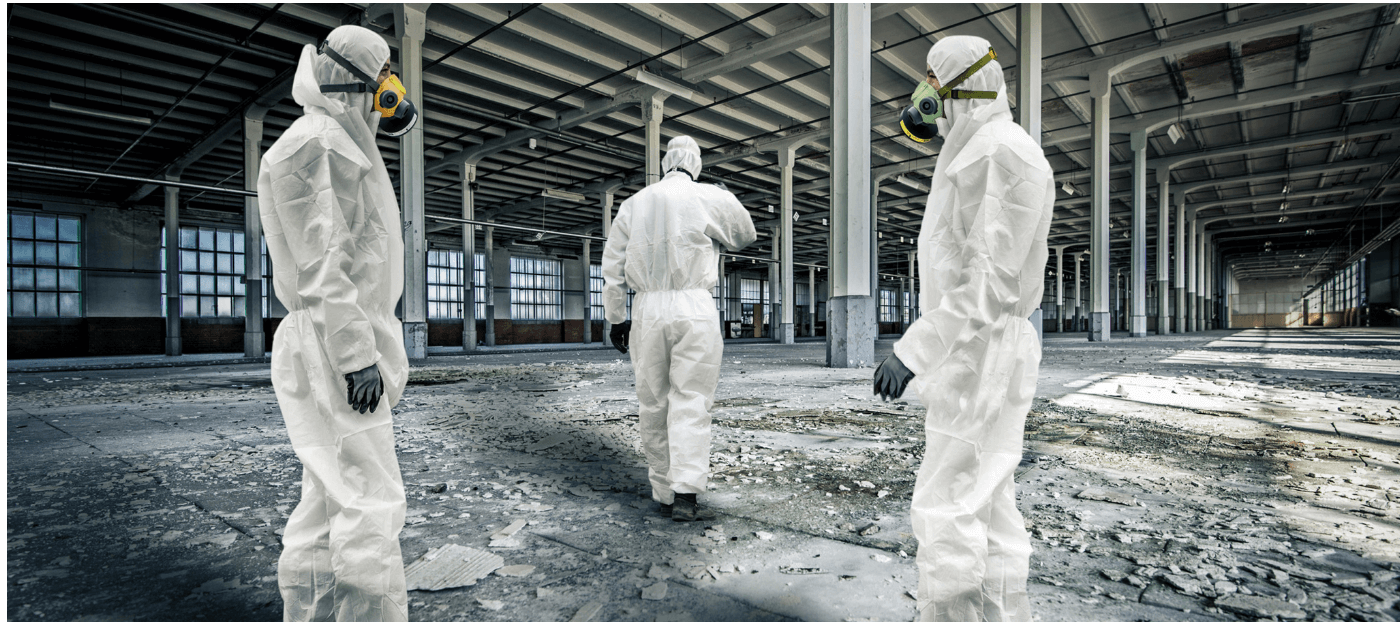 Drei Personen in Schutzanzügen und Atemschutzmasken gehen durch einen verlassenen, industriellen Raum mit beschädigtem Boden. Die Umgebung wirkt leer und unordentlich, was auf eine sanierungsbedürftige Situation hinweist.