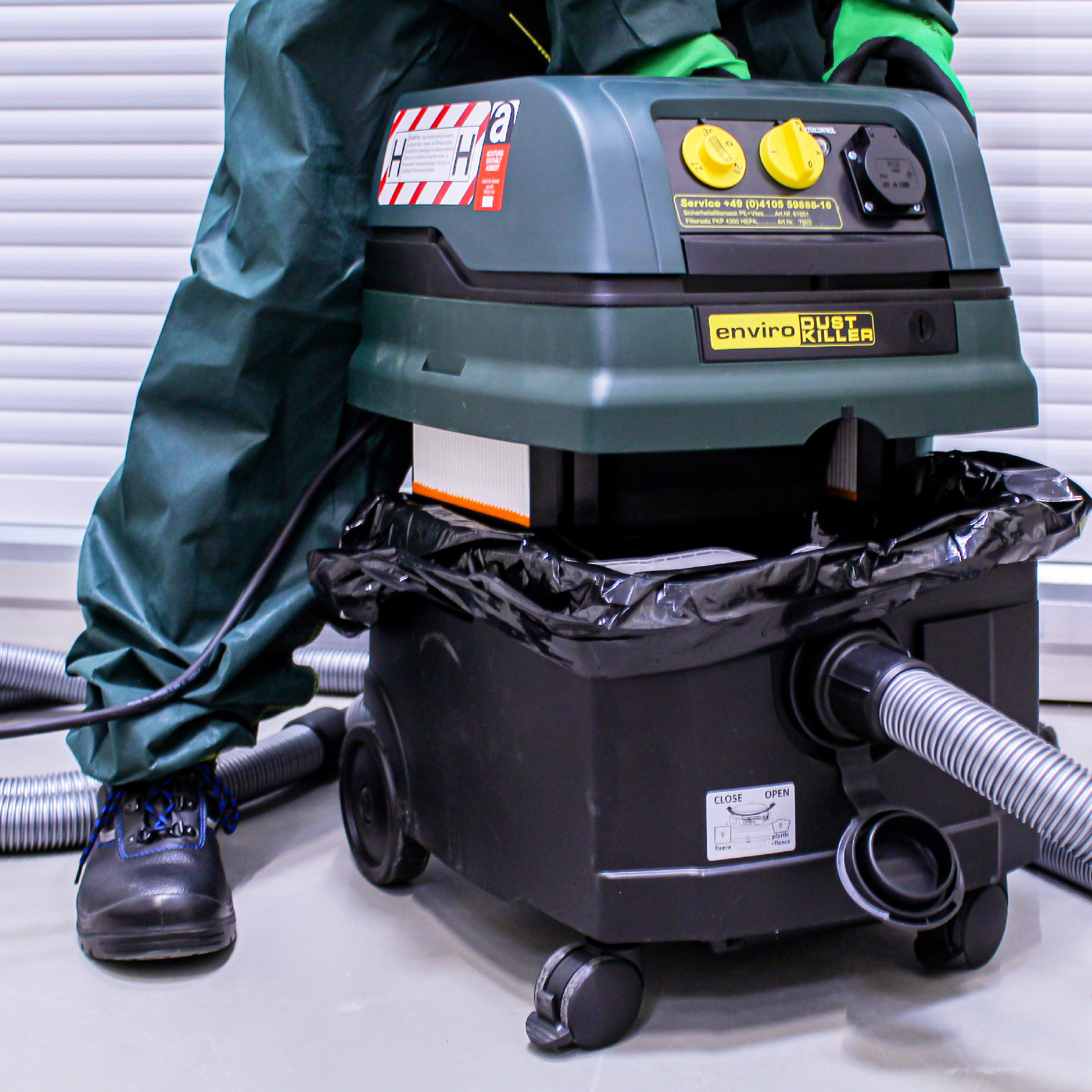 Ein grüner Industrie-Staubsauger steht auf dem Boden, teilwiese mit einem schwarzen Müllsack umgeben. Er hat zwei gelbe Knöpfe und einen Schlauch, der zu ihm führt. Eine Person in Schutzkleidung steht neben dem Staubsauger, bereit zur Benutzung.
