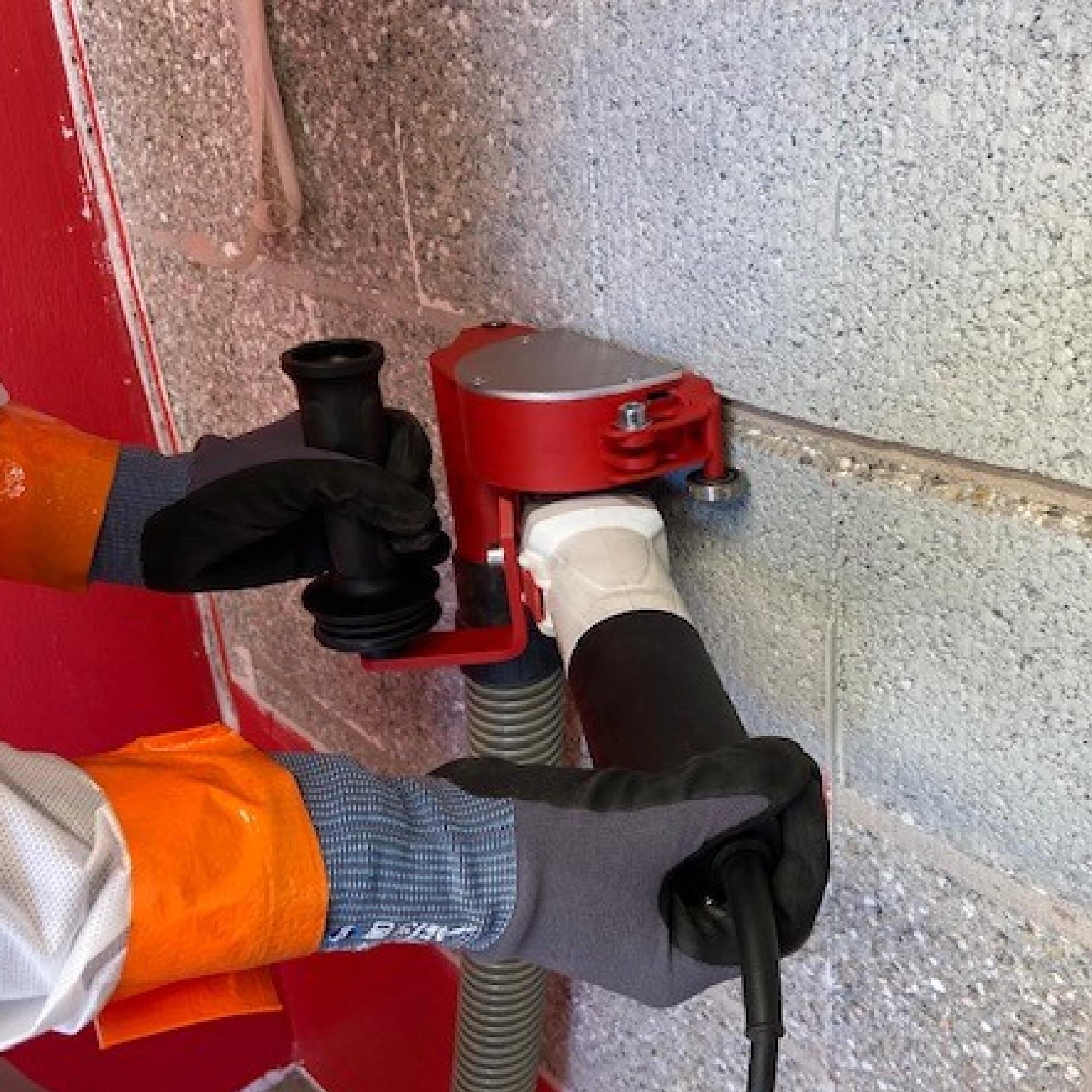 Eine Person trägt Handschuhe und hält mit beiden Händen ein rotes Gerät, das an einer Wand montiert ist. Das Gerät hat einen großen schwarzen Schlauch und einen weißen Anschluss. Die Wand ist aus grauen Steinen und hat eine rote Fläche.
